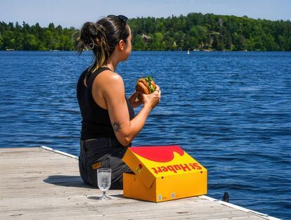 Woman sitting on a wooden dock by a lake, seen from behind, holding a St-Hubert burger. A large yellow St-Hubert box is placed next to her, along with a glass of water. The background shows a blue lake and forest under a sunny sky.