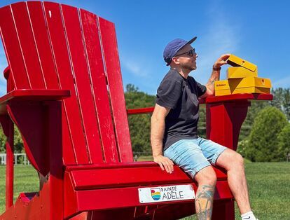 Un homme assis sur une immense chaise Adirondack rouge dans un parc ensoleillé à Sainte-Adèle, tenant des boîtes jaunes St-Hubert posées sur l’accoudoir. Il porte un t-shirt noir, des shorts en jeans, une casquette et des lunettes de soleil.