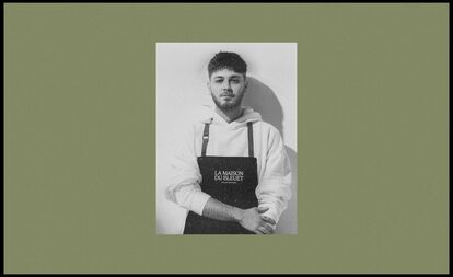 A black and white portrait of a person looking at the camera, wearing a dark apron over a light sweater. The apron clearly displays the 