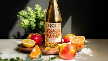 Bouteille de sangria La Dorada du Domaine Lafrance, étiquette colorée, entourée de pommes rouges, demi-citron jaune, demi-orange, quartier d’orange, planche en bois, arrière-plan noir, plante verte floue.