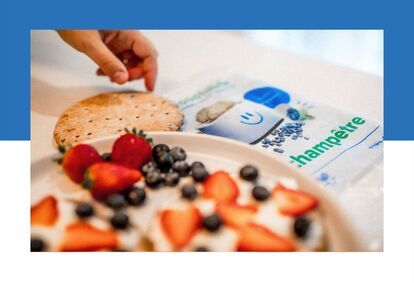 A hand picks up a bun next to “La Food Fabrik” brand packaging with a smiling face logo and the words “champêtre” (“country”). A plate of rolls garnished with strawberries and blueberries is visible in the foreground.