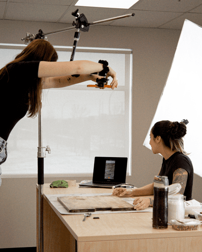 Deux personnes en séance photo-vidéo, une tenant une caméra au-dessus d’une table, l’autre assise préparant le décor, ordinateur portable ouvert en arrière-plan, ambiance de studio créatif avec éclairage professionnel.
