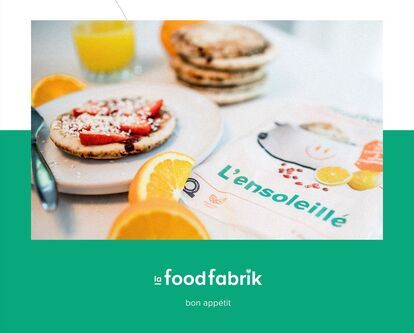 Une assiette avec un pain déjeuner garnie de fraises, de noix de coco râpée et de sirop, accompagnée d'un verre de jus d'orange et de tranches d'orange. À côté, un emballage alimentaire de la marque
