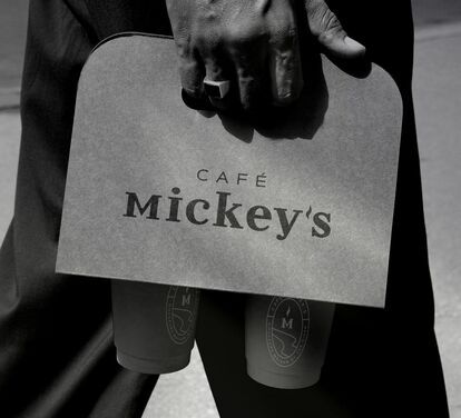 Black and white photo of a person holding a Café Mickey's coffee bag with two cups. The logo and name of the café are visible on the bag and cups.