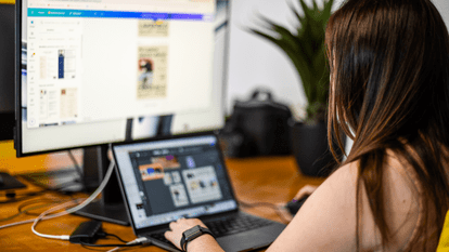 Personne de dos devant deux écrans, un portable et un moniteur affichant des maquettes graphiques, main sur la souris concentrée sur une interface de design, plante en arrière-plan sur un bureau en bois.