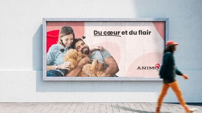 Panneau publicitaire installé sur le mur blanc d'une ruelle en brique grise pale. Le panneau est de deux personnes assises et entrelacés, avec un chien beige à poile frisé dans leur bras. Le logo ANIMO et le texte