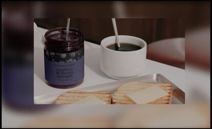 An open jar of wild blueberry jam from La Maison du Bleuet with a spoon inside, accompanied by a cup of coffee and two buttered toasts on a white plate.