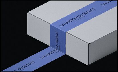 A closed rectangular white box adorned with a wide blue ribbon wrapped diagonally around it. The ribbon features the name 