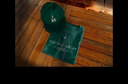 Dark green cap and t-shirt with the Café Mickey's logo and slogan, placed on a wooden floor.