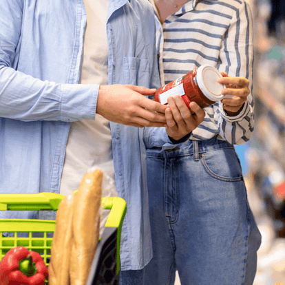 Couple en épicerie, tenant un pot de sauce rouge et consultant l’étiquette, panier avec baguettes et poivron rouge, vêtements décontractés, arrière-plan flou d’allée de magasin, vérification des informations nutritionnelles.