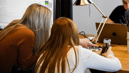 Deux personnes de dos concentrées devant un écran et un téléphone, doigt pointant une image affichée, ambiance de travail collaboratif sur une table en bois, lampe de bureau et ordinateur portable en arrière-plan.